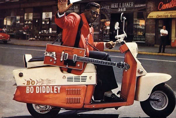 Bo Diddley montando un scooter personalizado diseñado para parecerse a su característica guitarra rectangular